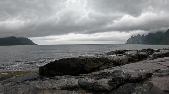Static ultra wide view of smooth granite shore and Okshornan peaks across ocean in Senja Norway