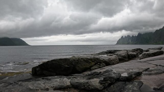 Static ultra wide view of gentle waves splashing on Tungeneset granite rocks with Okshornan peaks in Senja Norway