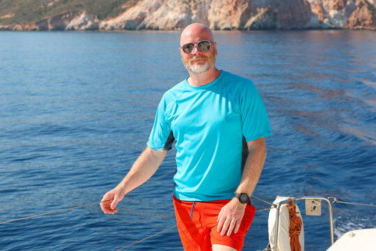 Bald man with beard wearing sunglasses on a sailboat touring the island of Milos in Greece. Aegean sea water behind him.