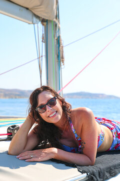 A smiling woman wearing sunglasses and a floral swimsuit relaxes on a sailboat near Milos, surrounded by calm blue water and bright Mediterranean sunlight.