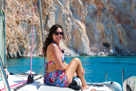 A smiling woman wearing sunglasses and a floral swimsuit relaxes on a sailboat near Milos, surrounded by calm blue water and bright Mediterranean sunlight.
