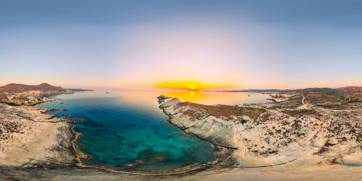 Sunrise over Mandrakia on Milos paints the sky in gold and orange hues above turquoise water and rugged coastal cliffs, revealing the tranquil beauty of the Aegean Sea.