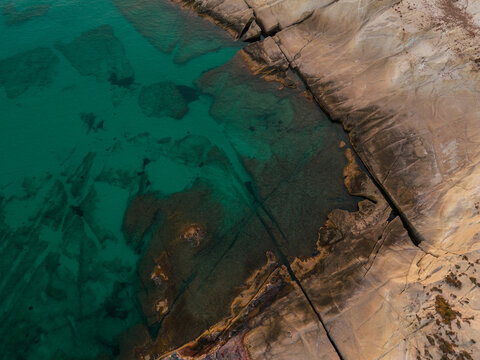 Aerial view of turquoise water and smooth rocky shoreline near Sarakiniko Beach on Milos, revealing unique geological patterns and the vivid colors of the Aegean Sea.