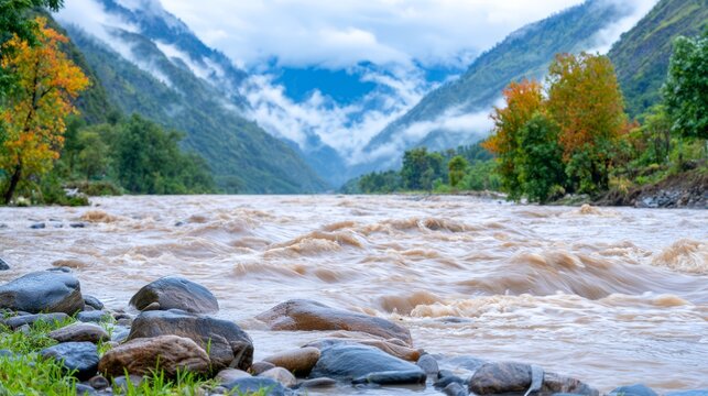 Severe flooding in a mountain valley, with a swollen muddy river rushing past rocky banks, misty forested slopes, and autumn trees under dramatic cloudy skies.
