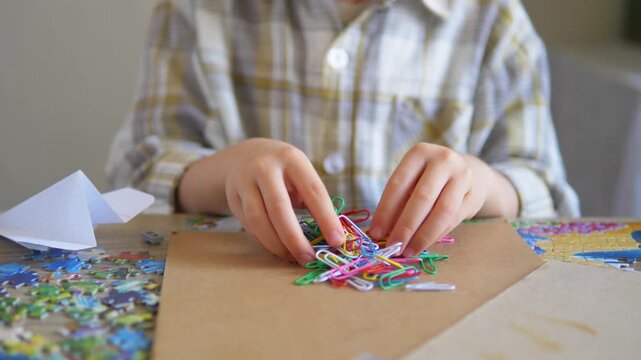 Close up of a child's hands playing with colorful paperclips next to a paper airplane on a messy desk