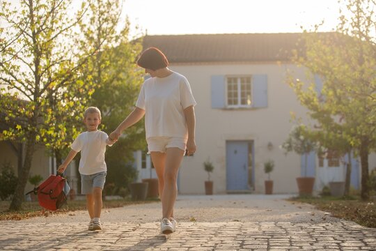 Mother and son walking hand in hand, in cobbled path house with blue shutters, with red backpack, cobblestones, from a eye-level perspective perspective, with white, blue accents.