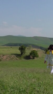 A mother and her little daughter in a white dress watching a colorful kite fly high in the blue sky on a green hill, slow motion vertical video.