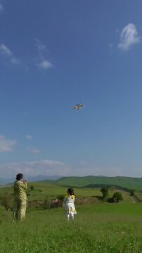 A mother and her little daughter in a white dress watching a colorful kite fly high in the blue sky on a green hill, slow motion vertical video.