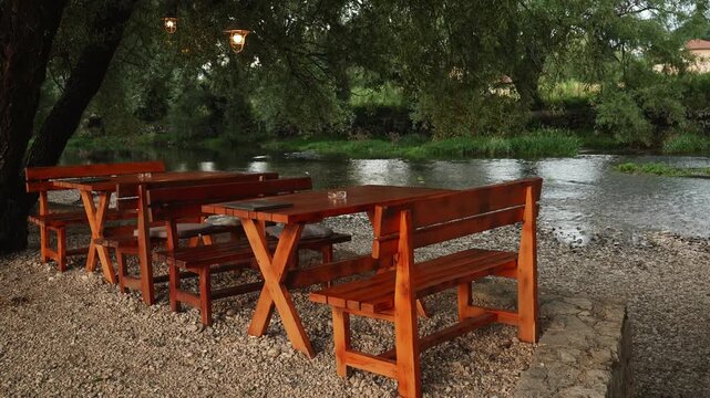 Cozy riverside dining creates a relaxing atmosphere for outdoor leisure and slow travel. Wooden tables and benches stand beneath trees beside a gently flowing river