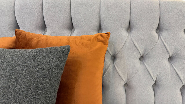 Close-up of gray tufted upholstered furniture with orange and dark gray decorative cushions, showcasing contrasting colors, soft fabrics and modern interior styling.