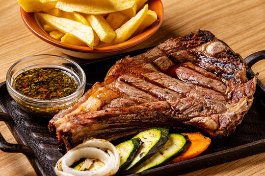 Grilled Ribeye Bone-In Steak Served on Cast Iron Skillet with French Fries, Grilled Vegetables, and Chimichurri Sauce