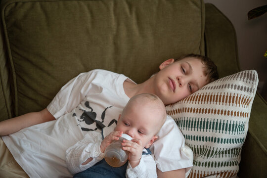 Older brother watches over baby sibling while younger child drinks from bottle resting in his arms on sofa at home. Sibling care, brotherhood, caregiving, responsibility, family bond, childhood