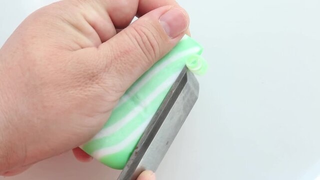 The hand grips the green striped soap tightly as the metal scissors slowly cut off a small piece, revealing the soft, pleasantly elastic texture in a slow, relaxing ASMR motion.