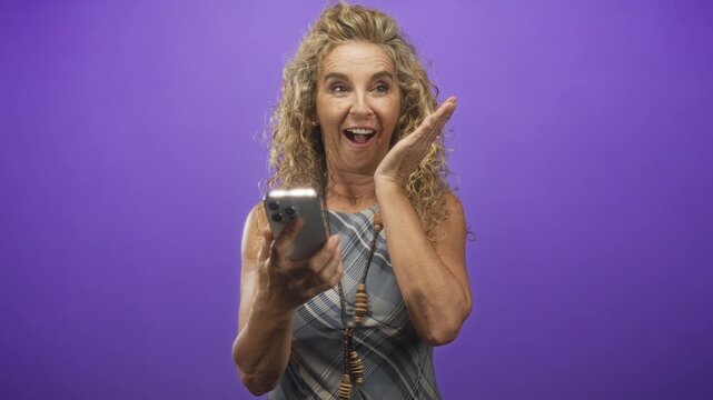 Woman middle age blonde holding smartphone, touching cheek and smiling while scrolling in purple studio; surprise connection.