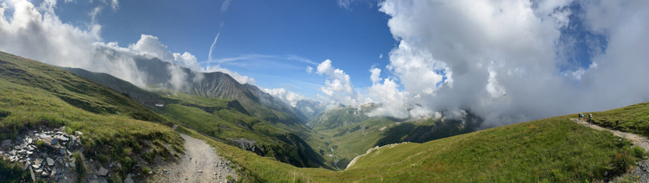 Alpine mountain pass panoramic view rocky peaks glacier green slopes hikers group dirt trail small tarn blue sky white clouds summer landscape wilderness valley