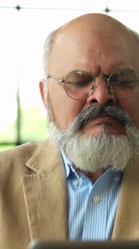 Indian Asian elderly businessman with a beard, dressed in a coat, seated in a modern office with glass walls,  working on a laptop, analyzing a business proposal 