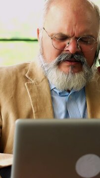 Indian Asian elderly businessman with a beard, wearing a suit, seated in a chair in a modern office, using a laptop while speaking on the phone about business matters, explaining about work