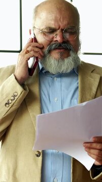 Indian Asian elderly businessman with a beard and suit,with documents in hand ,sitting in a revolving chair while discussing important business matters on the phone 