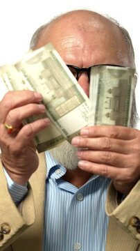Indian Asian senior businessman standing against a white backdrop,counting Indian currency ,showing gestures by fist of hands and smile