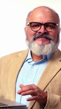 An Indian Asian senior businessman with a beard stands against a white background wearing a suit, holding a laptop in one hand and  pointing in a different direction, suggesting an important idea