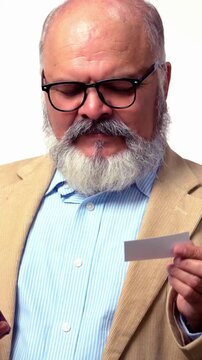 An Indian Asian old businessman with beard,in a suit, standing against a white background,is  holding a phone and using a debit card for online shopping