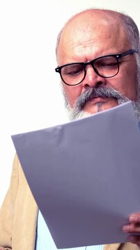 An Indian Asian elderly businessman with a beard and suit is standing against a white background crosschecking documents or calculations on a smartphone with a worried expressions