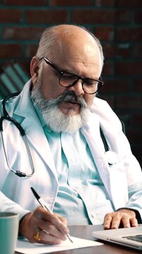 Indian Asian elderly doctor with a beard, wearing an apron and stethoscope, is having an online consultation on his laptop in a modern clinic, listening patiently to the patient and taking notes