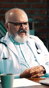 Indian Asian elderly doctor with a beard, wearing an apron and stethoscope, is having an online consultation on his laptop in a modern clinic