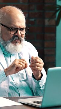 Indian Asian senior doctor with beard in white coat, stethoscope around neck, conducting virtual consultation  on a laptop in a modern clinic, explaining  tratment details