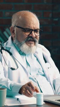 Indian Asian elderly doctor with beard, in white coat and stethoscope, explaining treatment and showing medicine on a laptop during online consultation at modern clinic