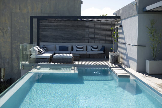 Rectangular turquoise pool is rippling on rooftop deck, with rattan sectional under slatted pergola