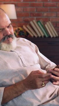 Indian Asian senior businessman with beard relaxing on sofa at night, using cellphone, struggling to get up because of back pain, then sits back in discomfort at home