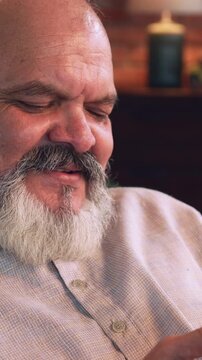Indian Asian elderly businessman with beard relaxing on sofa at night at home, using cellphone, suddenly experiences chest pain chest