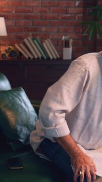 Indian Asian elderly businessman with beard relaxing on sofa at night at home, using cellphone, suddenly feels severe knee pain while trying to get up, showing a painful expression on his face