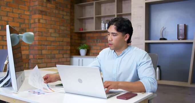 Asian man in blue shirt noticing color charts, checking between monitor and laptop while typing
