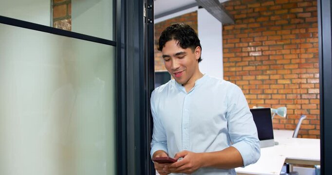 Glancing down at smartphone, man leaning on frosted glass scrolling, replying to messages in office