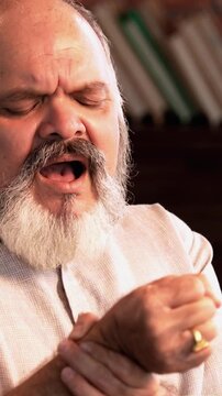 Indian Asian elderly businessman with a beard is sitting on a sofa in his modern home, feeling severe wrist pain while trying to move his wrist, showing a painful expression on his face