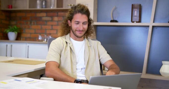 Man wearing beige shirt reacting to funny laptop message, smiling then laughing at office desk