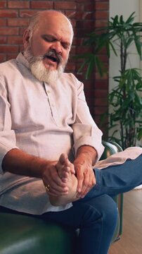 Indian Asian elderly businessman with a beard is sitting on a sofa in his modern home, experiencing severe ankle pain while trying to exercise his ankle, showing a painful expression on his face