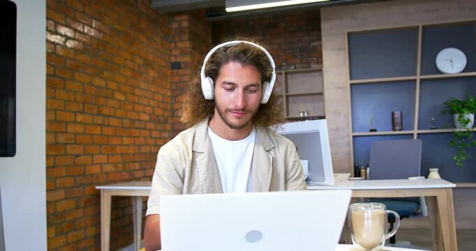 Man sitting at office desk, typing on laptop, responding to call via headset, smiling with mug
