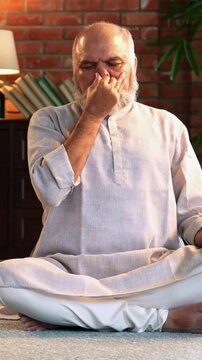 An Indian Asian elderly businessman with a beard is sitting comfortably on the floor of his modern home, wearing traditional Indian clothes and doing breathing exercises for health and  mental peace