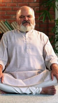 An Indian Asian elderly businessman with a beard is sitting on the floor in comfortable modern home, dressed in Indian clothes and doing breathing exercises to for his health and  peace of mind