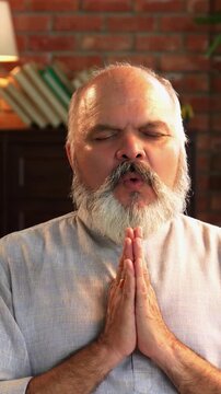 An Indian Asian elderly dressed in traditional Indian clothes and is chanting 'OM' with his hands folded and eyes closed, in a peaceful environment