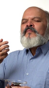 An Indian Asian elderly businessman with a beard is happily enjoying gulabjamun from a glass bowl with eyes closed , expressing great pleasure with hand gestures 