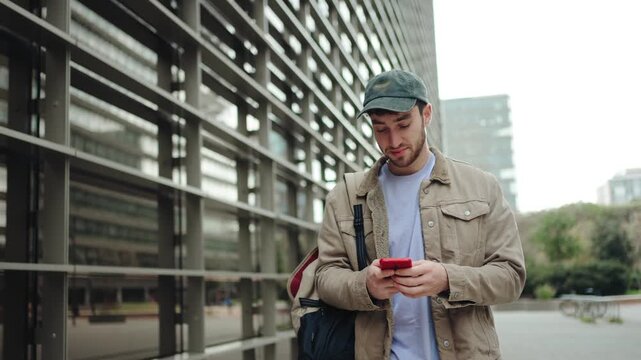 Young man with casual cap using mobile phone app in front of college campus building
