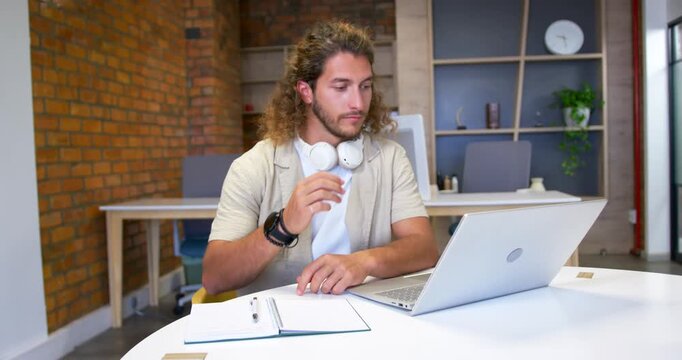 Man sitting at round white table in coworking office, working on silver laptop and rubbing temple