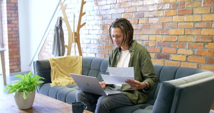 African American man reviewing papers and typing on laptop on blue sofa working remotely
