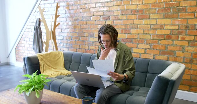 African American man sitting on tufted sofa at home reviewing pages after opening laptop for work