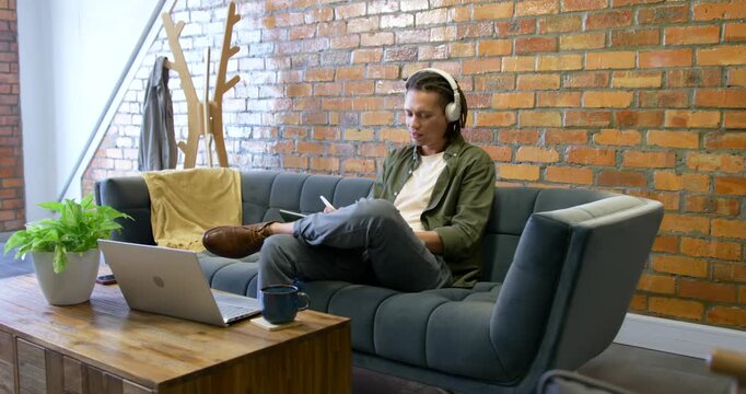 African American man with headset glancing left writing in notebook checking laptop working at home