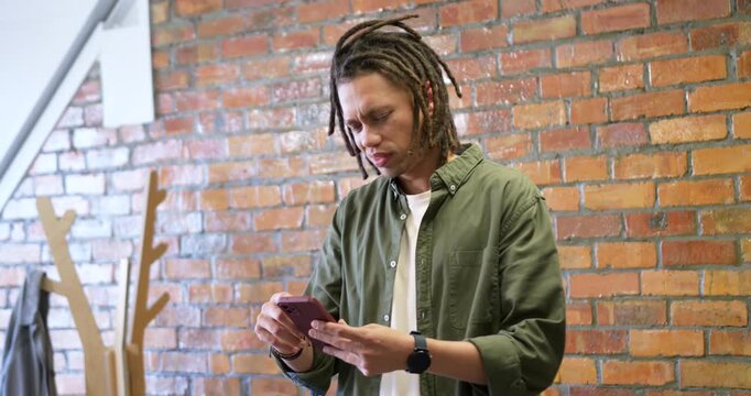 African American man checking pink phone, tapping, swiping, reacting to text in loft in olive shirt